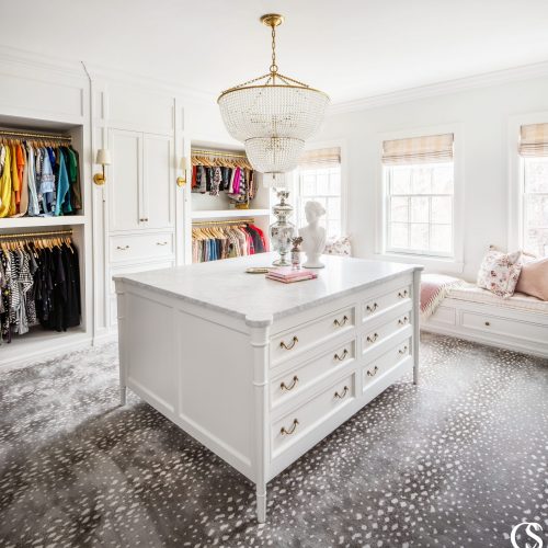 This light and airy custom closet design stays grounded with deep gold accents, a heavy marble countertop on the central built in cabinet, and a fashionable printed carpet.