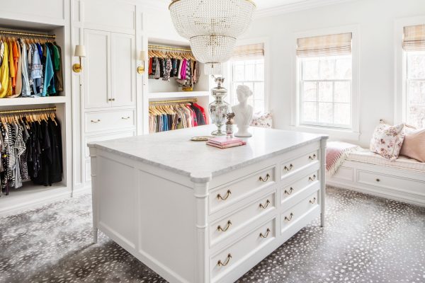 This light and airy custom closet design stays grounded with deep gold accents, a heavy marble countertop on the central built in cabinet, and a fashionable printed carpet.